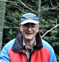 Photo of man with hat, glasses and blue and red jacket with forest in background